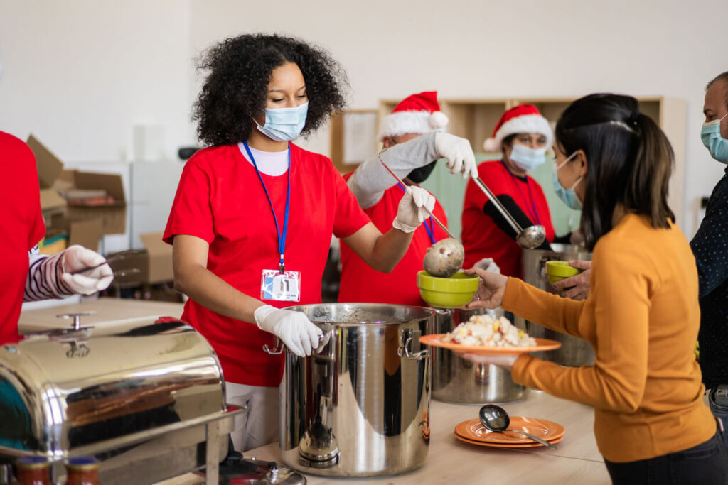 Multi-ethnic, mixed age group of volunteers work together at food bank during the Christmas season wearing protective masks and gloves, and cheery red t-shirts with Santa hats
