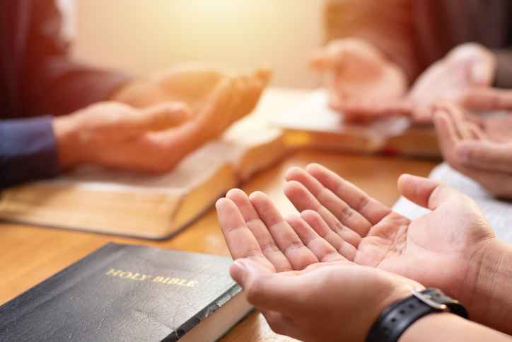 Power of hope or love, christian devotion background, bible study group concept. A group of people are gathered around a table with their hands outstretched in prayer over a Holy Bible.